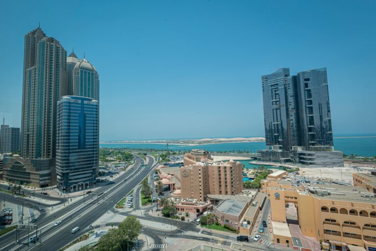 From above of contemporary city with skyscrapers and multistory buildings modern highway roads under blue cloudless sky near ocean
